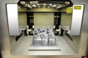 The view into the interior of the 3D printer shows the first printed thermostat covers, which are still connected to the work platform. After removal of the platform and support structure, the aluminium/silicon metallic powder is removed by suction, sieved, cleaned and ecologically fed back into the recycling system.